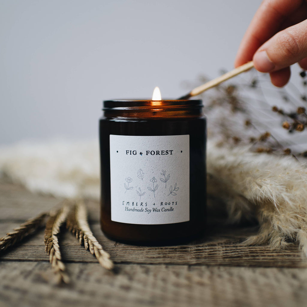 Candle labeled 'Fig & Forest' being lit with a wooden wick, on a textured surface with decorative elements.