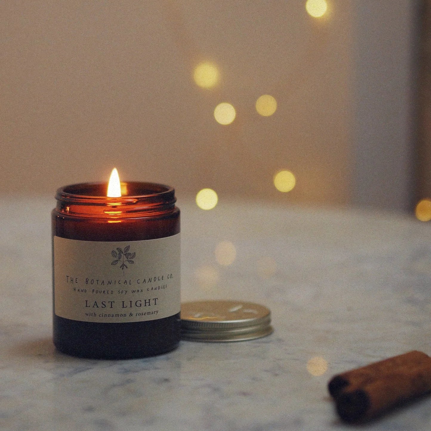 A lit amber glass jar candle labeled 'The Botanical Candle Co. Last Light' sits on a light marble surface with its metal lid beside it and warm string lights blurred in the background.