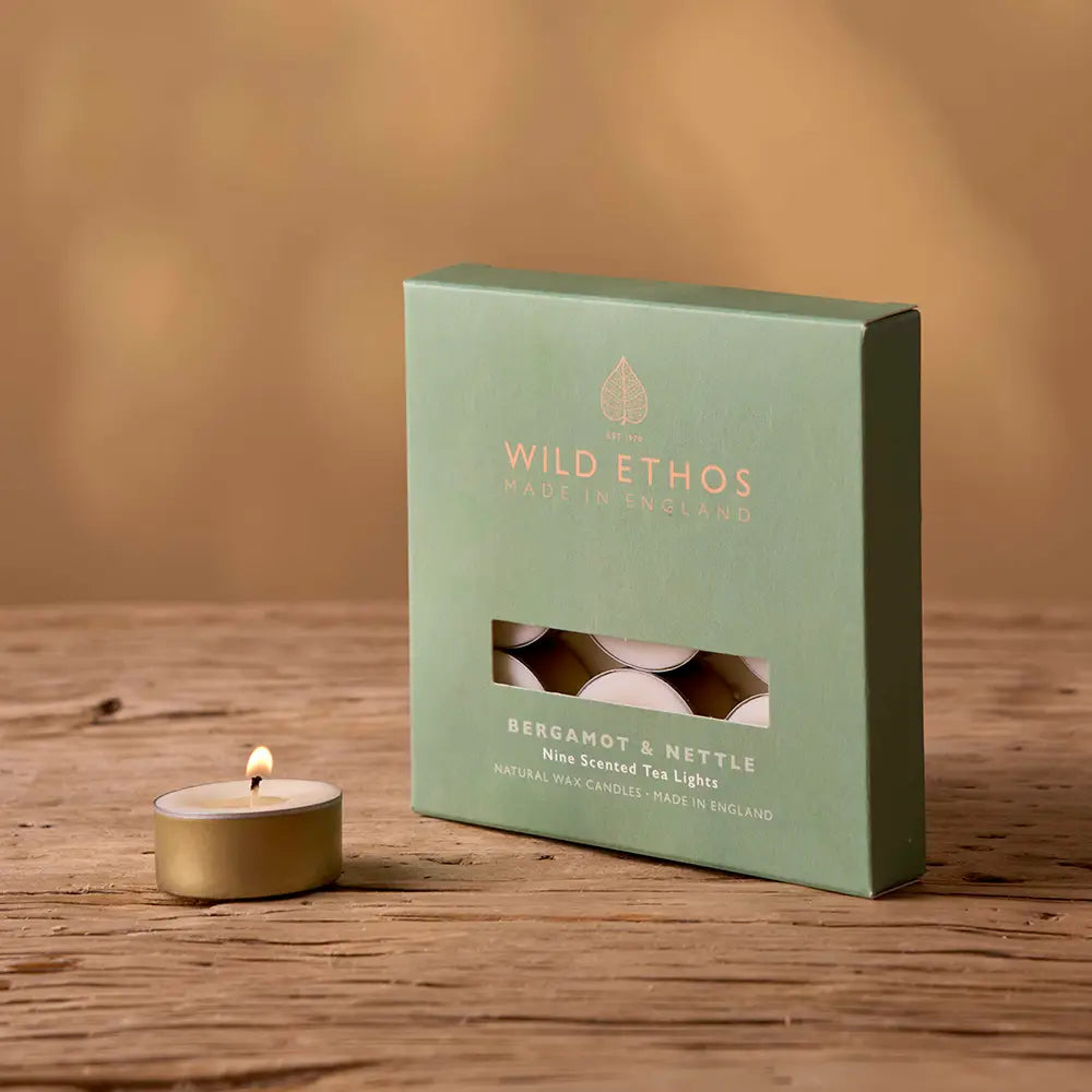 Green box of Wild Ethos tea lights on a wooden surface with a lit candle beside it.