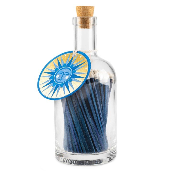 A glass bottle of navy matches, with a cork stopper and a tag around the neck featuring a gold sun illustration. 