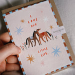 A hand holds a square greeting card with a kraft envelope behind it. The card has a cream background with a brown and blue striped border, an illustration of two horses, and the text 'I LOVE OUR LITTLE LIFE.