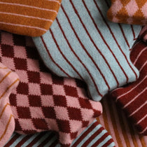 A close-up arrangement of hot water bottles in warm tones, including striped and diamond-patterned pieces in rust orange, dusty pink, burgundy red, and pale blue.