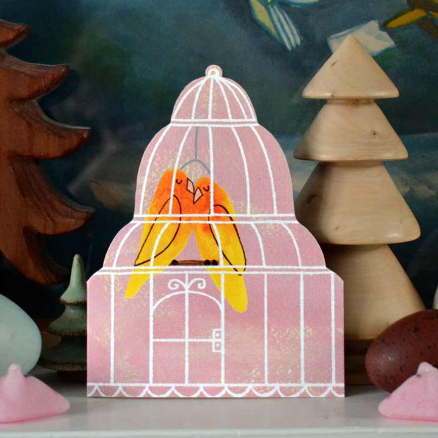 A die-cut card in the shape of a bird cage, created by Mortlake Papers, with two yellow/orange lovebirds perched inside.