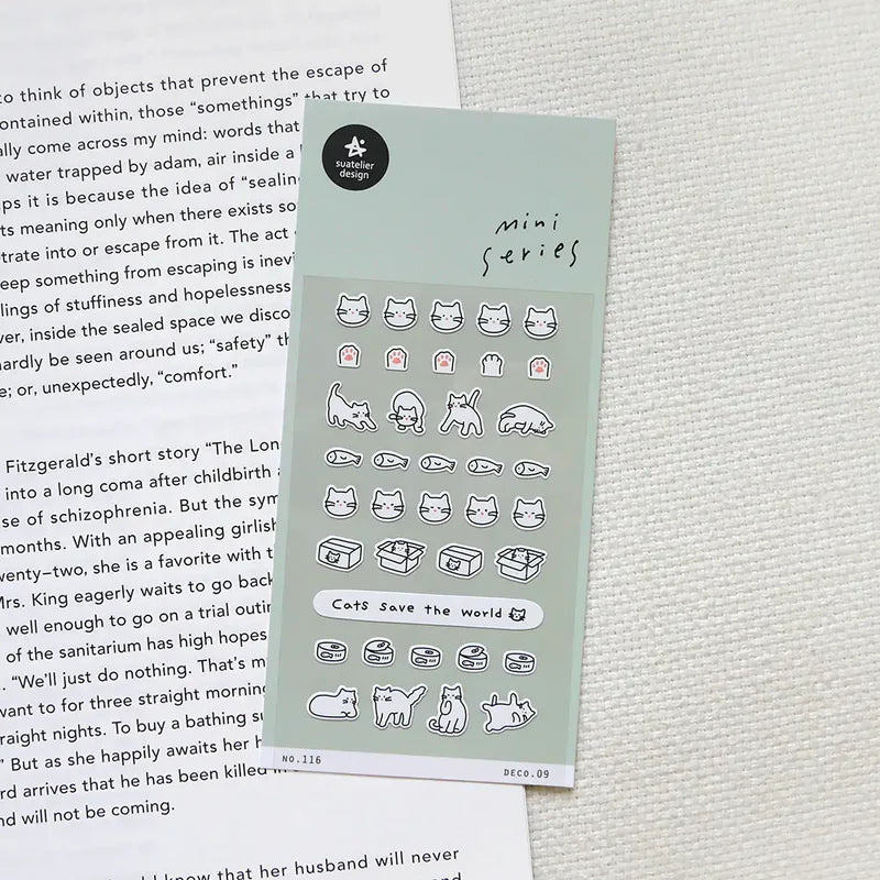 Sheet of stickers with various cat designs on a textured surface next to a book.